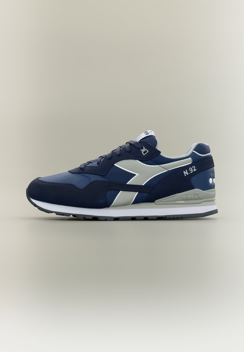 Ναυτικό μπλε και γκρι αθλητικό παπούτσι με κορδόνια, λωρίδα με την επωνυμία στο πλάι και λευκή σόλα, φαίνεται από την εξωτερική πλευρά σε ουδέτερο φόντο.