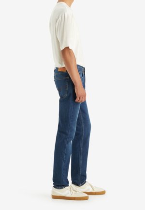 Homme portant une chemise blanche à manches courtes, un jean bleu et des baskets blanches, debout de profil devant un fond uni.