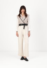 Gilet en maille beige avec des bordures noires sur une chemise blanche, associé à un pantalon ample crème et des chaussures noires. La ceinture ajoute un élément de structure.
