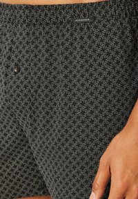 Shorts noirs avec un motif géométrique blanc, taille élastique, détail boutonné et texture de tissu lisse. Étiquette de marque visible à la taille.