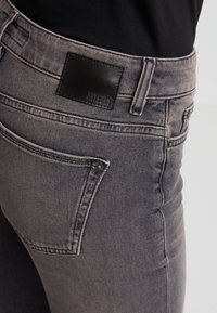 Vaqueros de mezclilla gris con un corte slim, bolsillo trasero y una etiqueta de cuero negro en la cintura, que exhiben una textura suave y un lavado sutil.