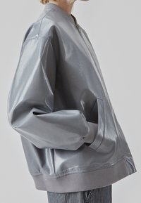 Veste bomber grise et brillante avec des poignets et un ourlet côtelés, dotée d'une fermeture éclair et de poches latérales. Texture lisse et coupe décontractée.