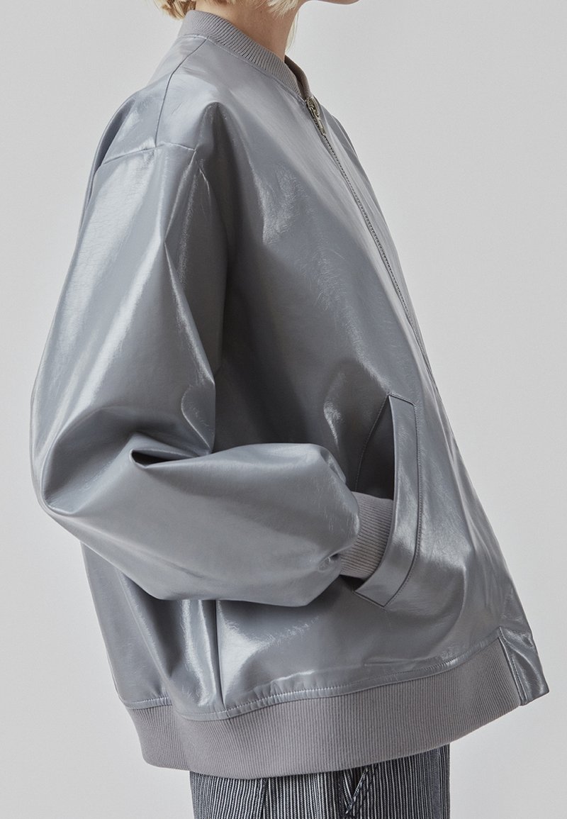 Veste bomber grise et brillante avec des poignets et un ourlet côtelés, dotée d'une fermeture éclair et de poches latérales. Texture lisse et coupe décontractée.