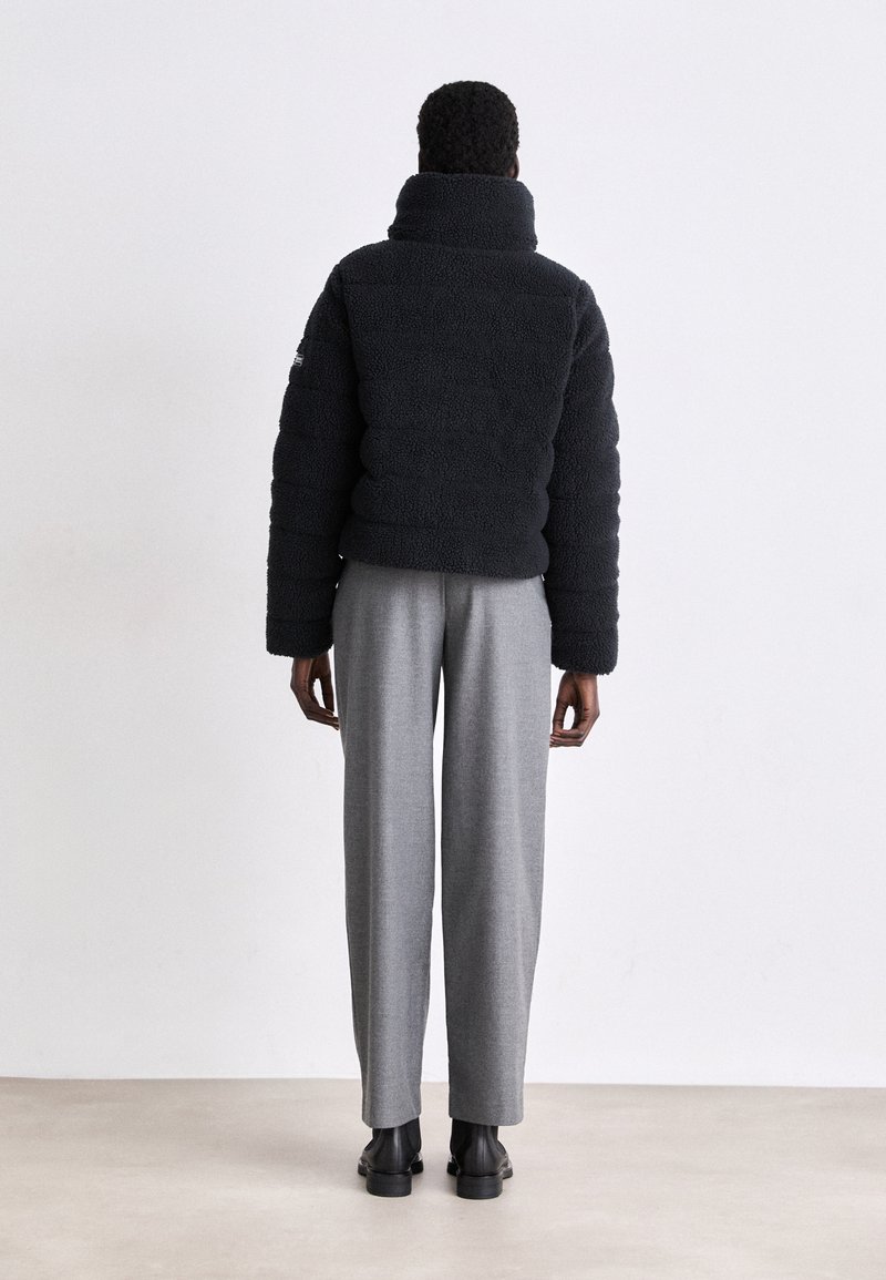 Giacca puffer nera in pile con collo alto e texture a coste, indossata con pantaloni grigi su misura e stivali neri alla caviglia.