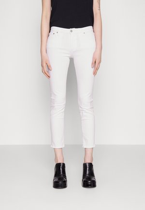 Personne portant un jean blanc ajusté coupé au-dessus des chevilles, un haut noir sans manches et des bottines à plateforme noires, debout devant un fond uni.