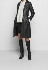 Black leather trench coat with a notched collar, button front, and side pockets, paired with a knitted light gray sweater, black mini skirt, and knee-high boots.