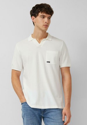 Junger Mann mit kurzen lockigen Haaren, der ein weißes Polohemd mit kurzen Ärmeln und Brusttasche sowie eine blaue Jeans trägt, vor einem einfarbigen Hintergrund stehend.