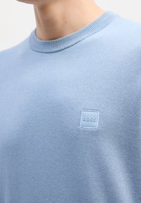 Pull bleu clair à col rond avec un petit logo carré « BOSS » sur la poitrine, porté par une personne.