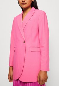 Růžový blazer vyrobený z hladkého materiálu, s jedním knoflíkem, klopami a dvěma předními kapsami. Přizpůsobený, jednobarevný design.