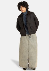 Veste en coton marron foncé avec un col et de grandes poches avant, portée sur un pull col roulé texturé bleu et une longue jupe en denim clair.