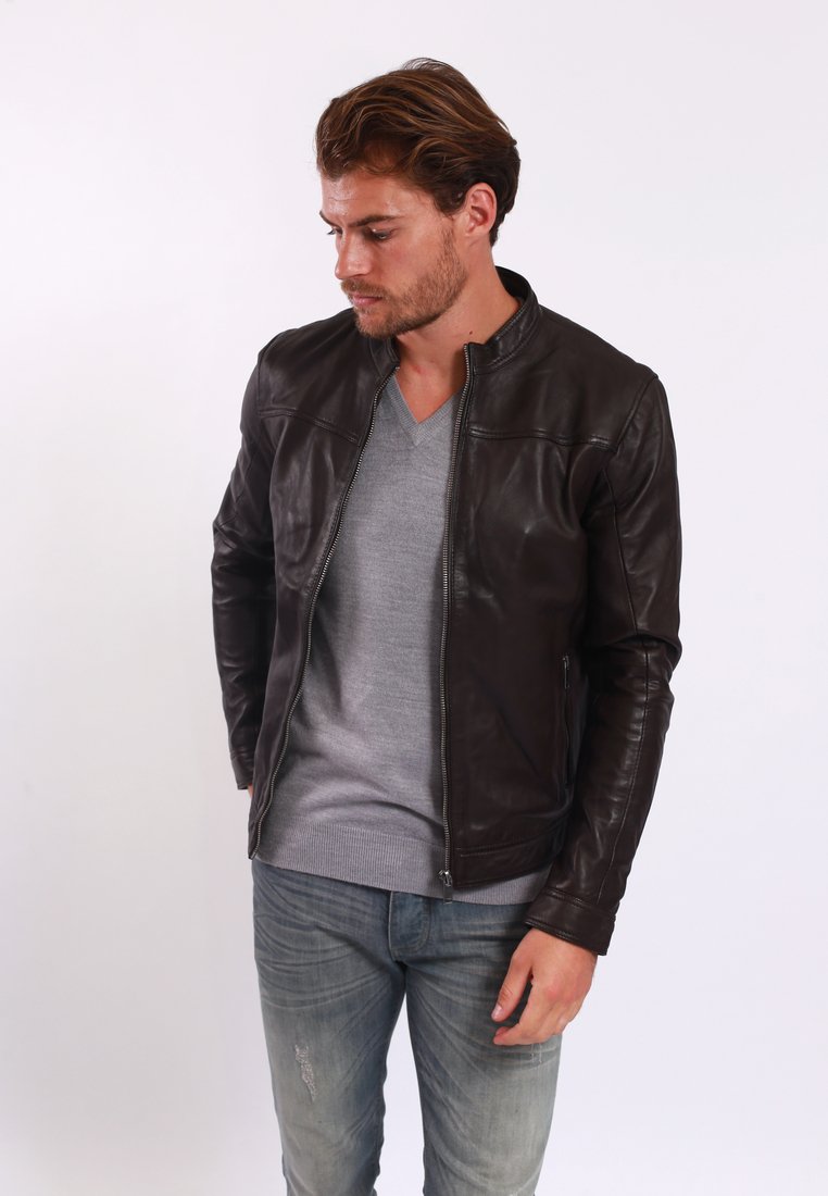 Veste en cuir marron avec un design ajusté, col montant et fermeture éclair à l'avant. Portée par-dessus un pull gris clair et assortie à un jean délavé.