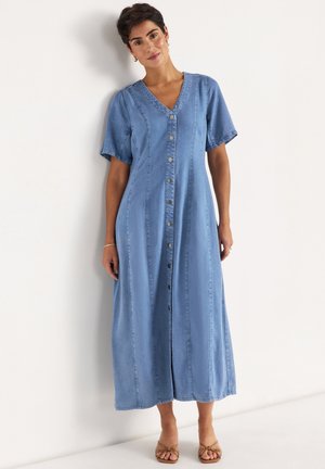 Kvinde med kort mørkt hår iført en lyseblå knap op midi denimkjole og brune åbne tåsandaler, stående mod en hvid væg.