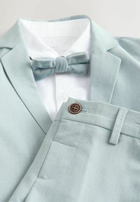 Svetlo modra suknjič in hlače s belo srajco ter ujemajočo se svetlo modro metuljčkom, položeni ravno na beli površini.