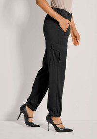 Schwarze tapered Jogginghose mit elastischem Bund und Seitentaschen, kombiniert mit glänzenden schwarzen High-Heels. Geschmeidige Textur und lässiges Design.