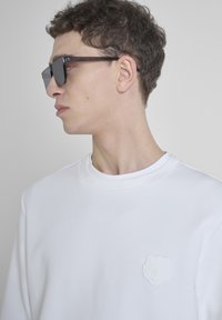 Sudadera blanca con cuello redondo, que presenta un parche de flor texturizada en el lado izquierdo del pecho. El modelo lleva unas gafas de sol rectangulares de color oscuro.