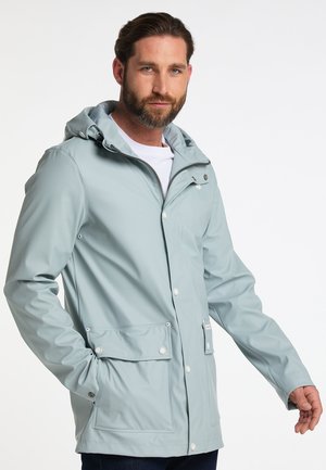 Mann trägt eine hellgraue, wasserdichte Kapuzenjacke mit Frontreißverschluss und Knöpfen, posiert mit den Händen in der Nähe der Taschen, weißer Hintergrund.