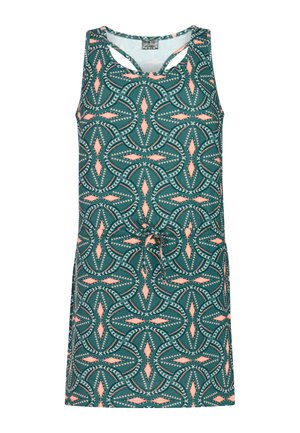 Meisje met lang bruin haar, een pet op, een mouwloze jurk met een patroon en witte slippers, staand tegen een effen lichte achtergrond.