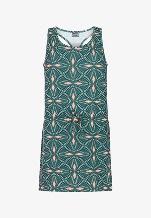 Meisje met lang bruin haar, een pet op, een mouwloze jurk met een patroon en witte slippers, staand tegen een effen lichte achtergrond.