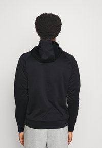 Haut à capuche noir au toucher doux et lisse, avec un capuchon ajustable et des manches raglan, associé à un pantalon de jogging gris.