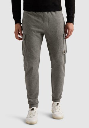 Homme portant un pantalon jogging cargo gris et des baskets blanches, debout devant un fond clair uni.