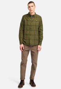 Groene en bruine geruite shirt met knoopsluiting, lange mouwen en kraag; gecombineerd met beige broek en bruine veterschoenen.