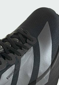 Black athletic shoe with textured fabric, reflective silver accents, mesh details, and flat laces. Features a streamlined, modern design.