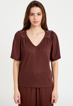 Ung kvinde med langt brunt hår iført en tekstureret brun V-hals bluse med blonde detaljer på skuldrene og korte ærmer, stående stille.