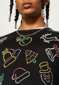 Schwarzer Strickpullover mit mehrfarbigem, festlichem Stickdesign, das Hüte, Glocken und Bäume zeigt. Ribbelkragen und sichtbare Kettenhalskette.