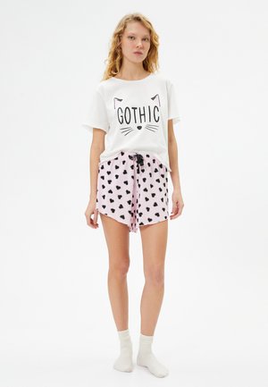 Witte katoenen t-shirt met "GOTHIC" print en kattengezicht ontwerp. Roze shorts met zwart hartpatroon, voorzien van een verstelbare taille.