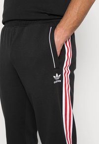 Čierne športové nohavice vyrobené z mäkkej tkaniny, s červenými a bielymi pruhmi na bokoch a logom Adidas na ľavom stehne.