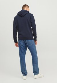 Dunkelblaues Kapuzensweatshirt mit gerippten Bündchen, kombiniert mit hellblauen, taillierten Jeans. Weiße Turnschuhe an den Füßen, Rückansicht gezeigt.