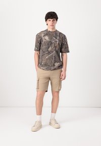 T-shirt à manches courtes camouflage dans des nuances de vert et de marron, associé à un short cargo beige avec poches latérales et une coupe décontractée.