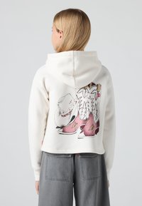 Sweatshirt cru cropped com uma gravação de botas de cowboy e um chapéu nas costas. Material macio com um corte descontraído. Calças cinzas parcialmente visíveis.
