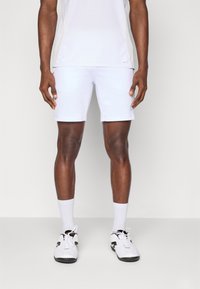 Λευκές αθλητικές βερμούδες με λείο υφή, που διαθέτουν μπλε λεπτομέρεια λογότυπου. Φοριούνται με λευκές κάλτσες και μαυρόασπρα αθλητικά παπούτσια.