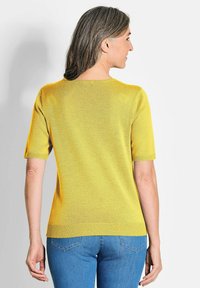 Kortebloesem gele trui van lichtgewicht breiwerk, met een ronde hals en ribgebreide zoom, gedragen met blauwe jeans. Achteraanzicht toont een gladde textuur.