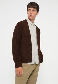 Brown knit cardigan with ribbed texture, featuring a button closure and two front pockets, worn over a light striped shirt and beige trousers.