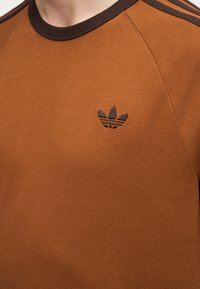 Καφέ μπλούζα Adidas με σκουρόχρωμη καφέ περιποίηση και τρεις ρίγες στον ώμο, με το κεντημένο λογότυπο Adidas trefoil στο στήθος.