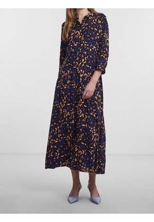 Frau trägt ein langärmeliges, knopfverschlossenes, knöchellanges schwarzes Kleid mit blauen und orangefarbenen abstrakten Mustern und hellviolette spitze Schuhe.