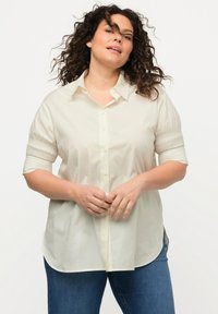 Helle Cremetön Button-Up-Bluse mit kurzen Puffärmeln, strukturierten Akzenten an den Ärmeln und einem abgerundeten Saum, kombiniert mit dunkelblauen Jeans.