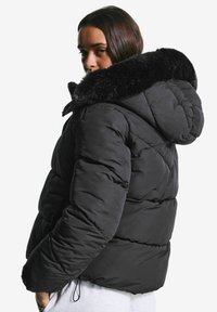 Vrouw in een zwarte gewatteerde jas met een grote met bont gevoerde capuchon, kijkt over haar schouder tegen een effen witte achtergrond.