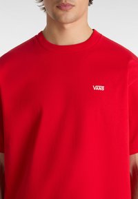Camiseta de algodón roja con cuello redondo, que presenta un pequeño logotipo bordado en blanco de "VANS" en el lado izquierdo del pecho. Tejido de peso medio con una textura suave.