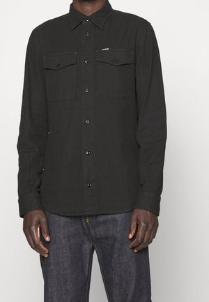 Czarna, długorękawowa koszula na guziki z dwoma kieszeniami na piersi, noszona nie wpuszczona w spodnie, na osobie stojącej w ciemnoniebieskich dżinsach.