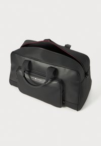 Black duffel bag made of smooth material, featuring dual handles, a front zipped pocket, and red zipper accents. Compact and rectangular shape.