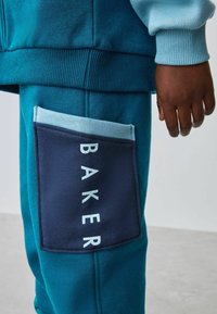 Los pantalones de chándal color azul verdoso tienen un gran bolsillo en parche azul marino con el texto blanco "BAKER". El material es suave, con una cintura y puños acanalados.