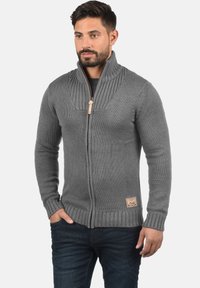 Cardigan grigio lavorato a maglia con zip e colletto a coste. Presenta cuciture testurizzate, una patch con logo sull'orlo inferiore e polsini aderenti.