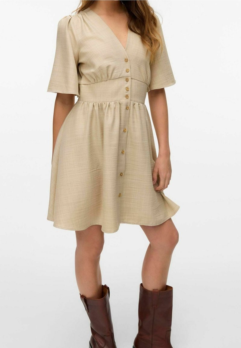 Femme portant une robe beige à col en V avec des boutons sur le devant, manches jusqu'au coude, et des bottes marron mi-mollet, debout avec une main tenant la jupe.