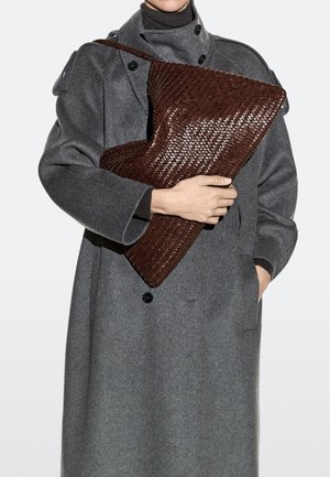 Person trägt einen grauen Wollmantel und hält eine große dunkelbraune gewebte Lederclutch vor weißem Hintergrund.