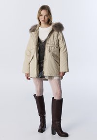 Beige gewatteerde jas met een met bont gevoerde capuchon, gedragen over een grijze top en een spijkerrok. Heeft visnetkousen en bruine kniehoge laarzen.