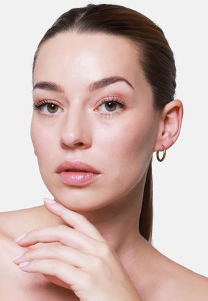 Boucles d'oreilles en or portées par une personne à la peau claire ; texture de peau lisse, maquillage naturel et ongles rose clair. L'arrière-plan est entièrement blanc.