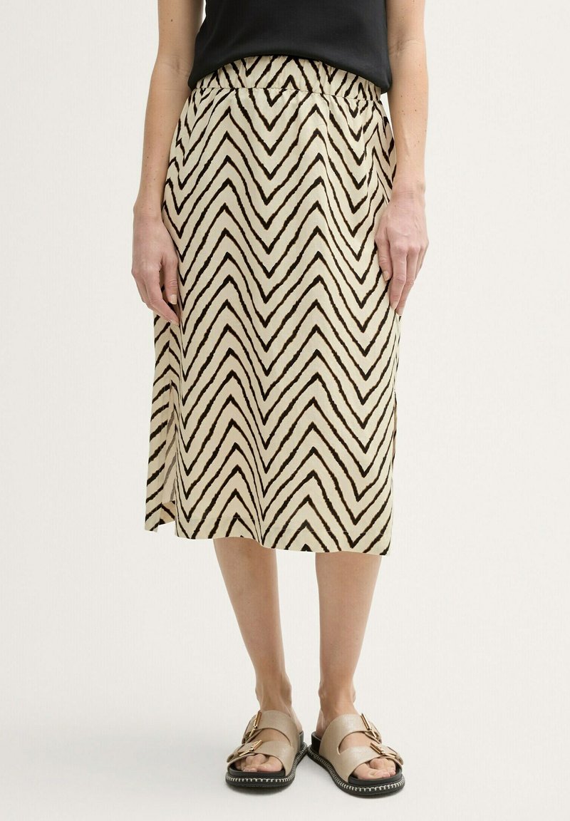 Jupe portefeuille crème avec un motif zigzag noir. Elle présente une taille élastique et une longueur mi-mollet. Portée avec des sandales beiges.